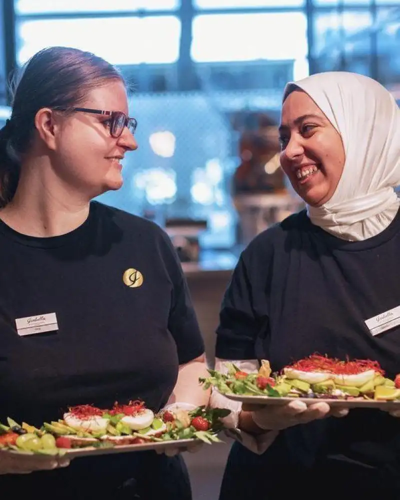 Zwei Frauen in einem Café. Beide halten einen Teller mit verschiedenen Speisen in ihren Händen und schauen sich an.