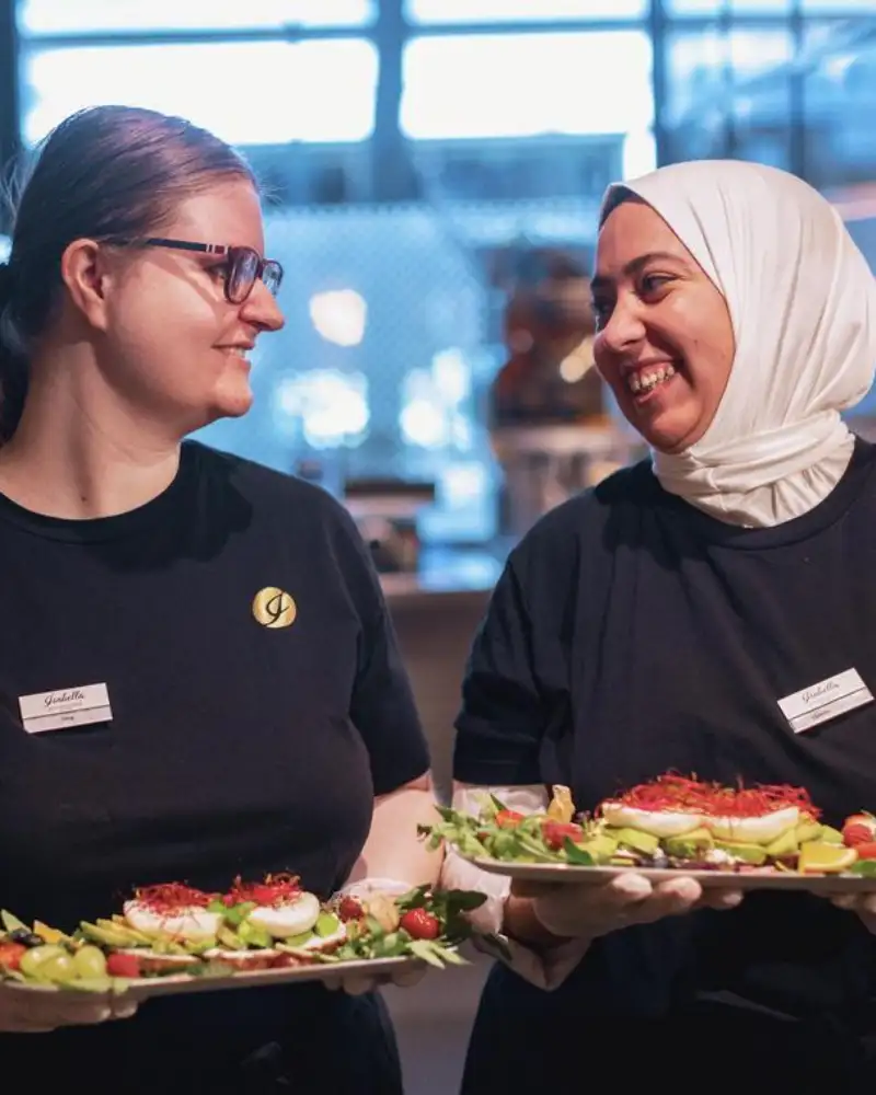 Zwei Frauen in einem Café. Beide halten Teller mit verschiedenen Speisen in ihren Händen und lächeln sich an.