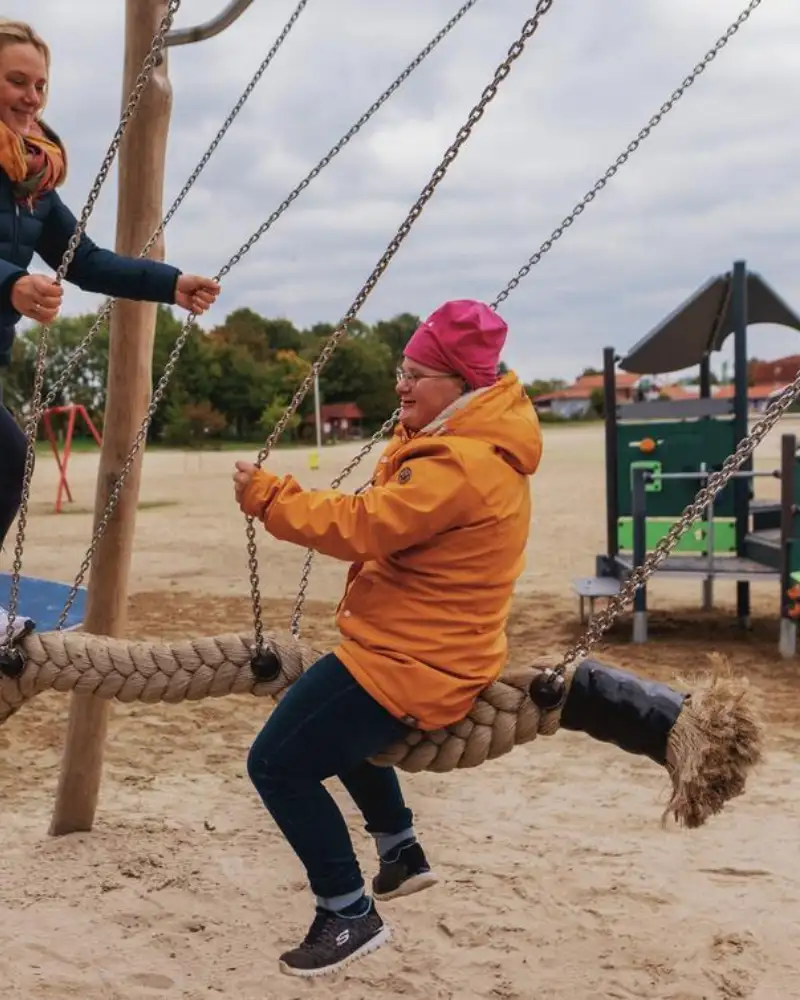 Eine junge Frau und eine Jugendliche auf einer Schaukel, die aus einem sehr dicken Tau gefertigt ist. Eine von ihnen steht, die andere sitzt auf dem Tau.