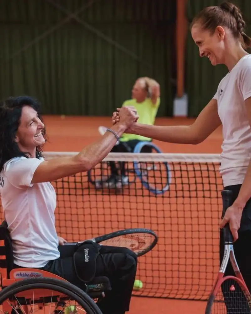 Auf dem Tennisplatz. Auf beiden Seiten des Netzes ist jeweils eine Person im Rollstuhl sitzend und eine Stehend. Die beiden vor dem Netz geben sich die Hand.