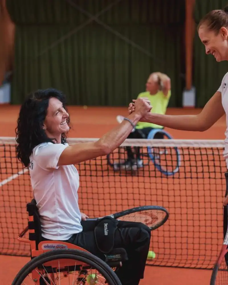 Auf dem Tennisplatz. Nach dem Spiel geben sich eine Tennisspielerin im Rollstuhl und eine stehende Tennisspielerin über das Netz hinweg die Hand. Im Hintergrund stehen zwei männliche Spieler. Einer von ihnen sitzt im Rollstuhl.