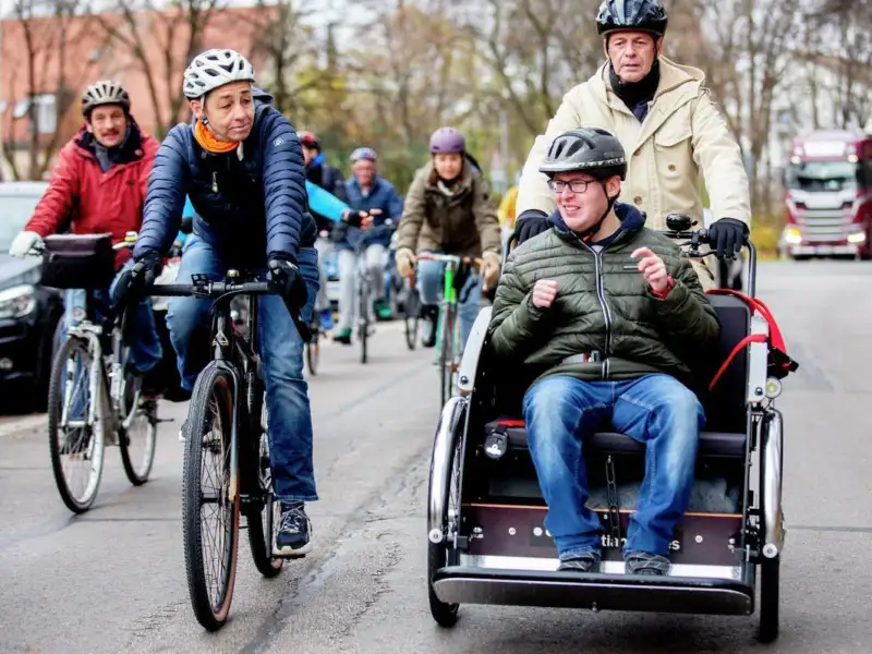 Vorneweg eine Fahrradrikscha mit einem Fahrer und einem Fahrgast. Daneben und dahinter ist eine Gruppe von Radlern unterwegs.