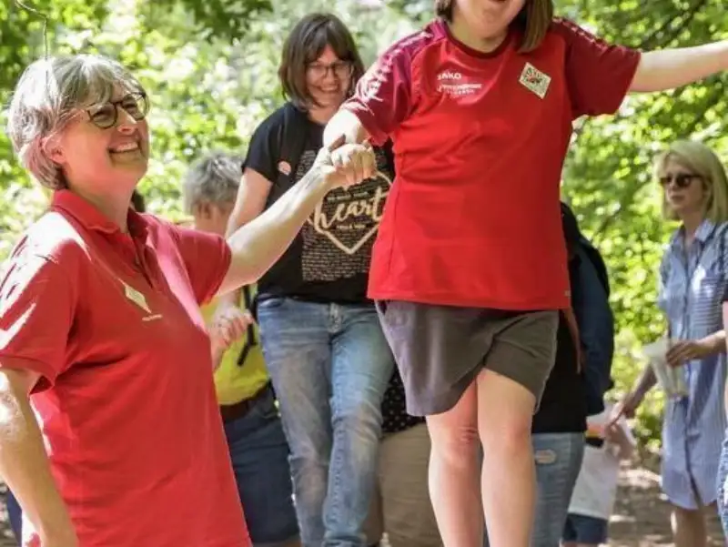 Ein Mädchen mit Down-Syndrom balanciert über einen Baumstamm. Dabei hält eine Frau ihre Hand. Dahinter folgt ein weiteres Mädchen. Weitere Personen stehen dabei.
