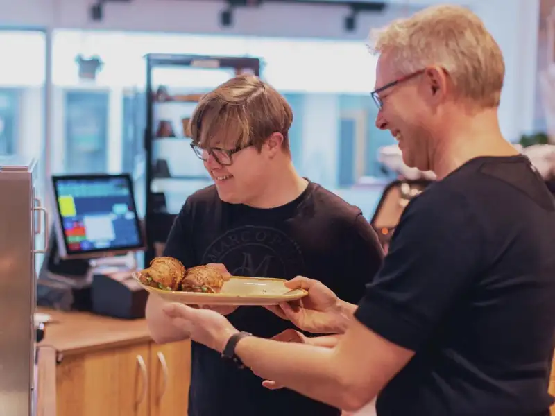 Zwei Erwachsene in einem Café. Der eine reicht dem anderen einen Teller mit Essen.