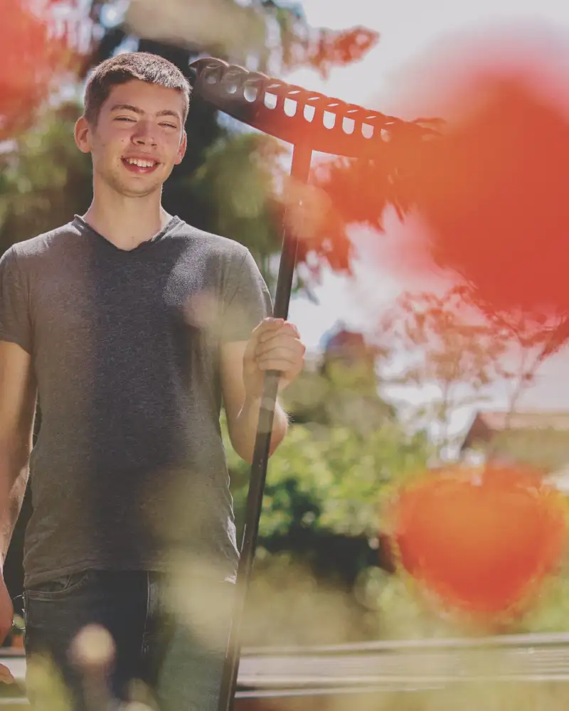 Ein Jugendlicher mit Harke im Grünen. Im Vordergrund sind orangefarbene lampionförmige Blumen im Bild.