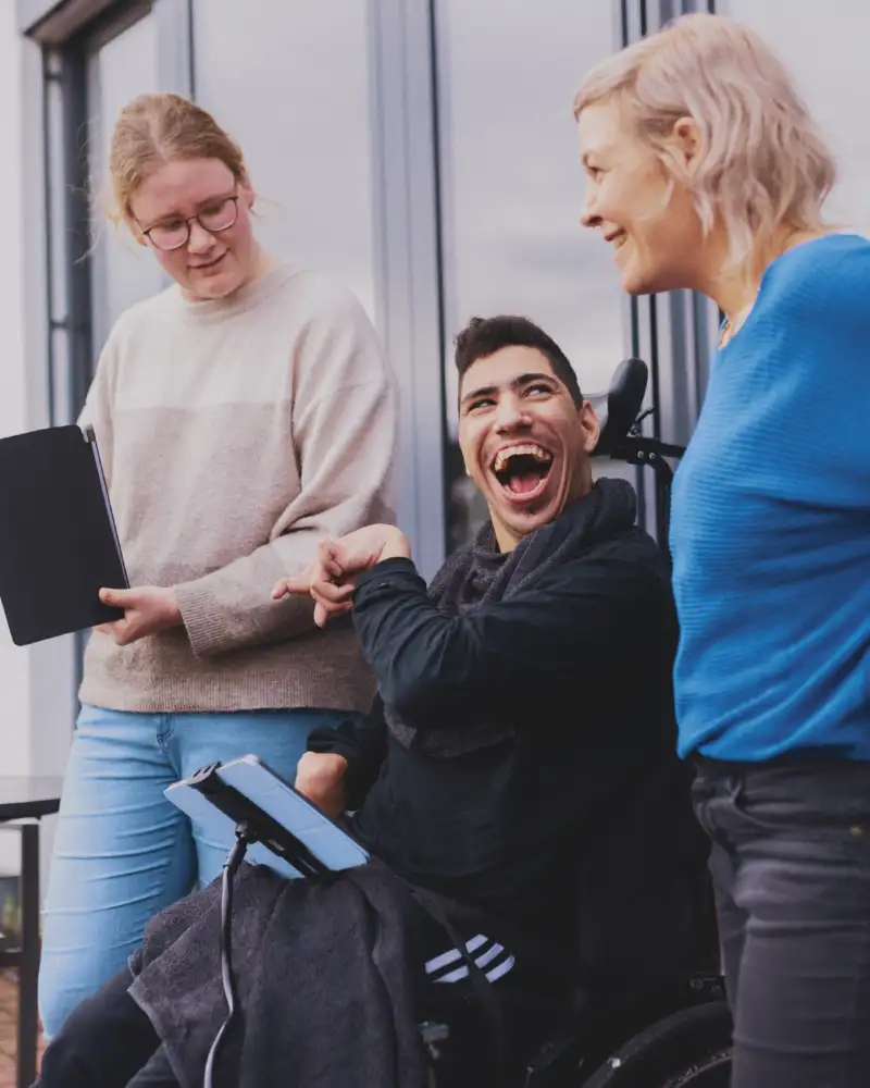 Ein junger Mann im Rollstuhl hält ein Tablet in der Hand und lacht in die Kamera. Er wird von zwei Frauen eingerahmt, von denen eine ebenfalls ein Tablet in der Hand hält.