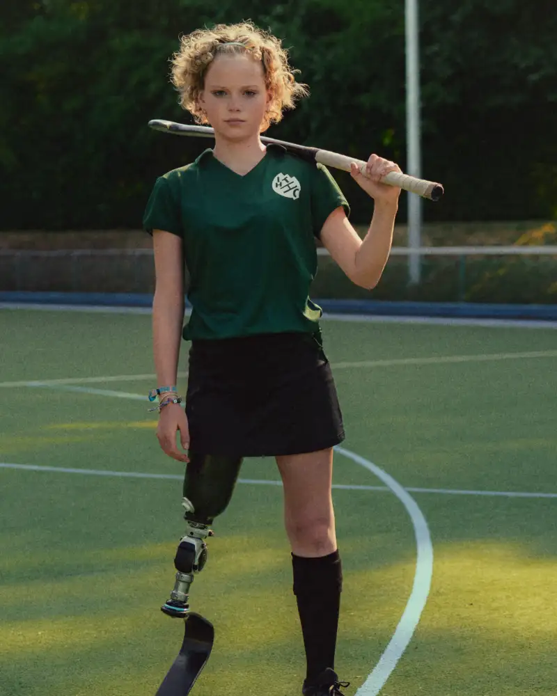 Eine Jugendliche mit Beinprothese trägt einen Hockeyschläger über der Schulter. Sie steht auf einem Rasenplatz. Hinter ihr steht ein Hockeytor.