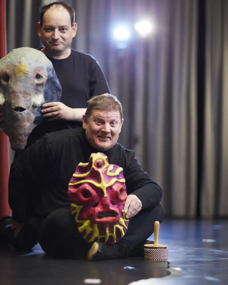 Zwei Männer auf einer Bühne. Einer hält eine Elefantenmaske in Händen, der andere die Maske eines Dämons.