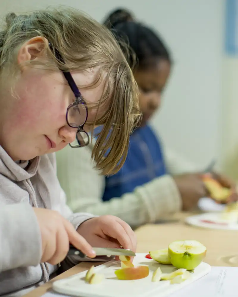 Ein Mädchen schneidet einen Apfel in kleine Stückchen. Mit ihr am Tisch sitzen weitere Kinder, die Obst schnippeln.