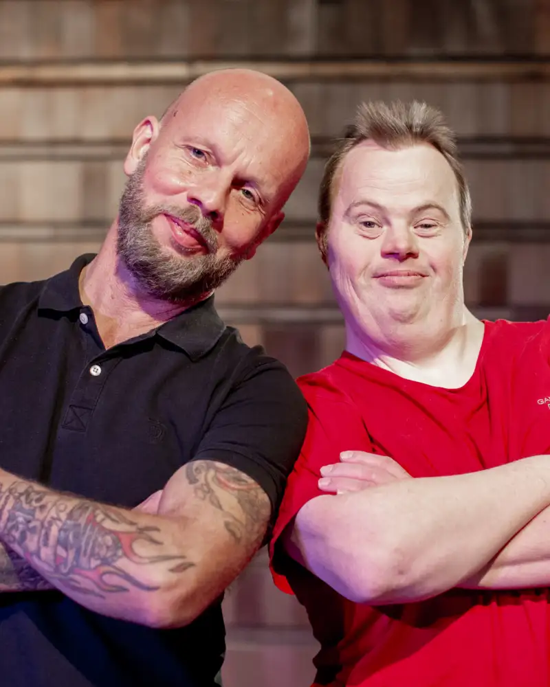 Zwei Männer verschränken die Arme und neigen ihre Köpfe zueinander. Einer von ihnen hat das Downsyndrom.