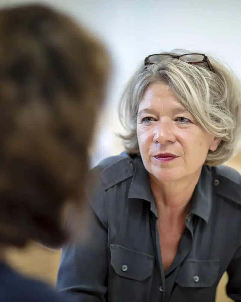 Eine mittelalte Frau, die die Brille in ihr graues Haar hochgesteckt trägt in einem Beratungsgespräch mit einer erwachsenen Person, die man nur angeschnitten von hinten sieht.