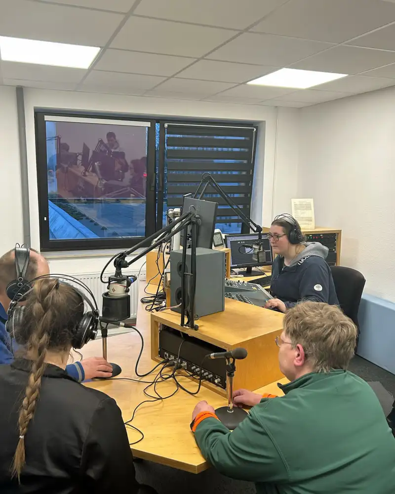Eine junge Frau mit Kopfhörern sitzt in einem Studio vor einem Mikrofon und einem Computer. Ihr gegenüber sitzen drei weitere Personen.