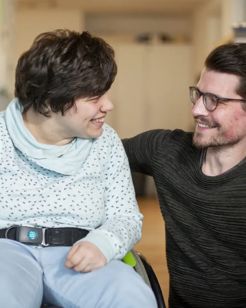Ein Mann hockt neben einer Frau im Rollstuhl. Die beiden lächeln sich an.
