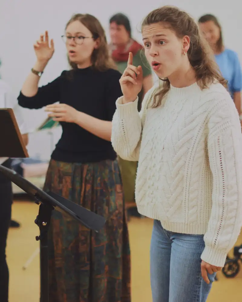 Drei junge Frauen singen. Vor ihnen steht ein Pult mit einem aufgeschlagenen Notenbuch.