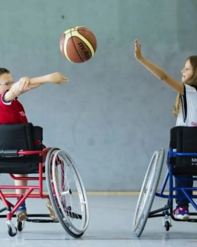 Ein Junge und ein Mädchen werfen sich einen Ball zu. Beide sitzen in Sportrollstühlen.