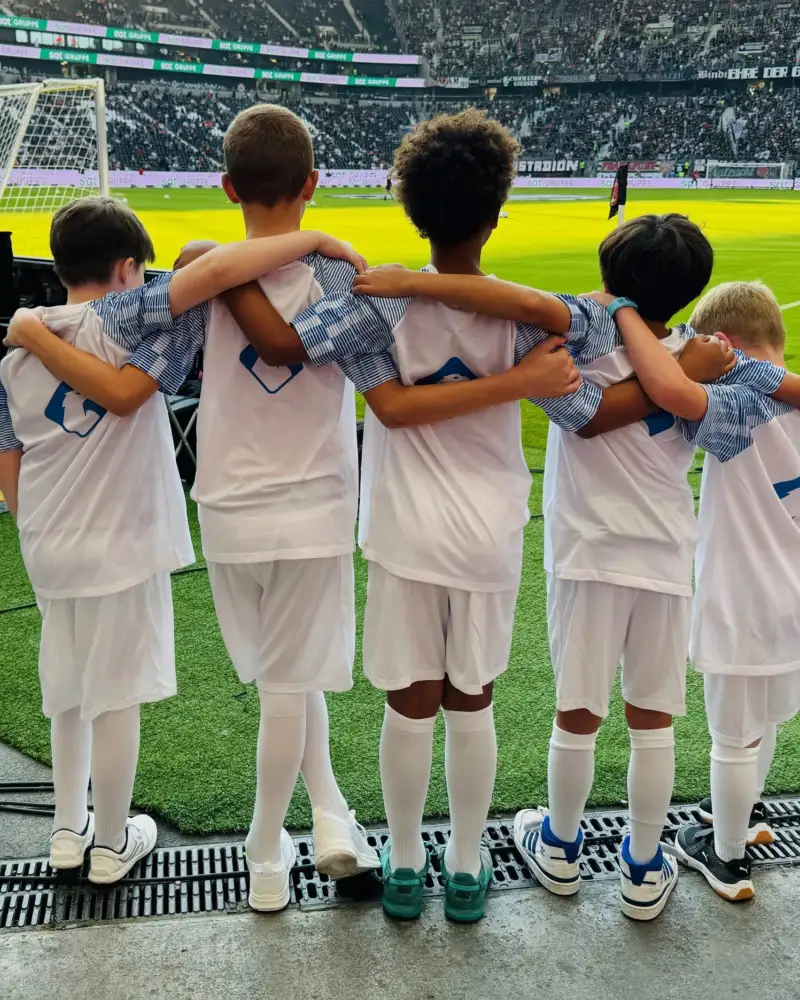 Fünf Jungen in weißen Trikots stehen am Rand eines Fußballfelds. Alle haben einander die Arme um ihre Schultern gelegt.