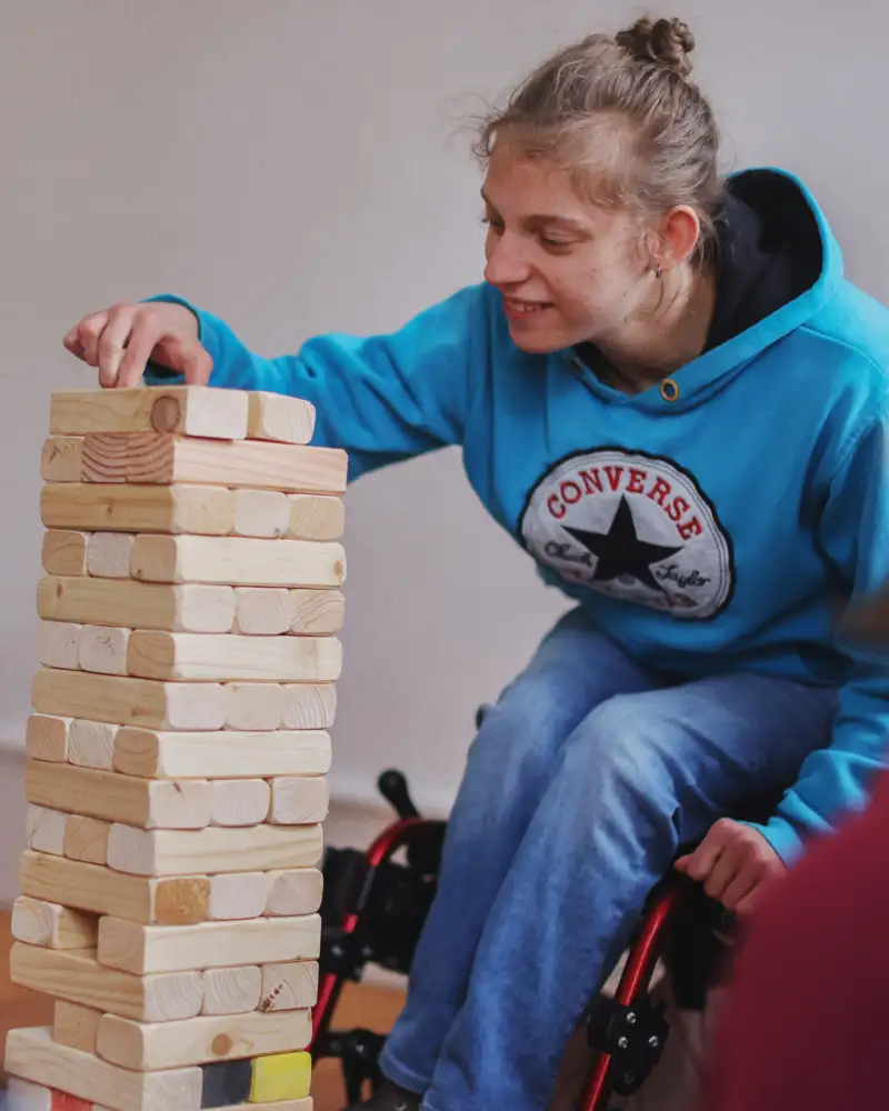 Eine Jugendliche sitzt im Rollstuhl und schichtet bearbeitete Holzstücke versetzt zu einem Turm auf.