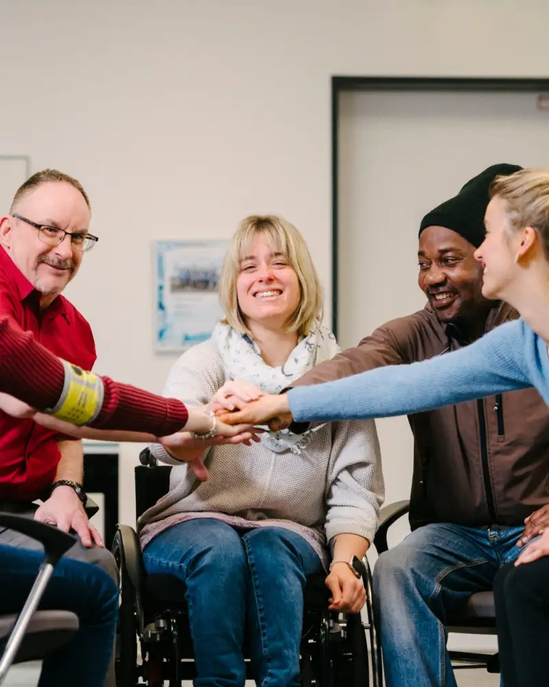 Zwei Frauen und zwei Männer sitzen in einem Büro in einem engen Kreis zusammen. Ihre rechten Arme sind ausgestreckt und ihre Hände treffen sich in der Mitte. Eine der Frauen sitzt im Rollstuhl.