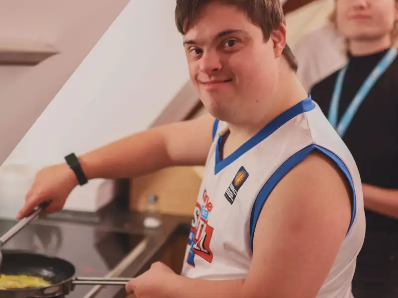 Ein junger Mann mit Downsyndrom kocht am Herd. Hinter ihm steht eine junge Frau.