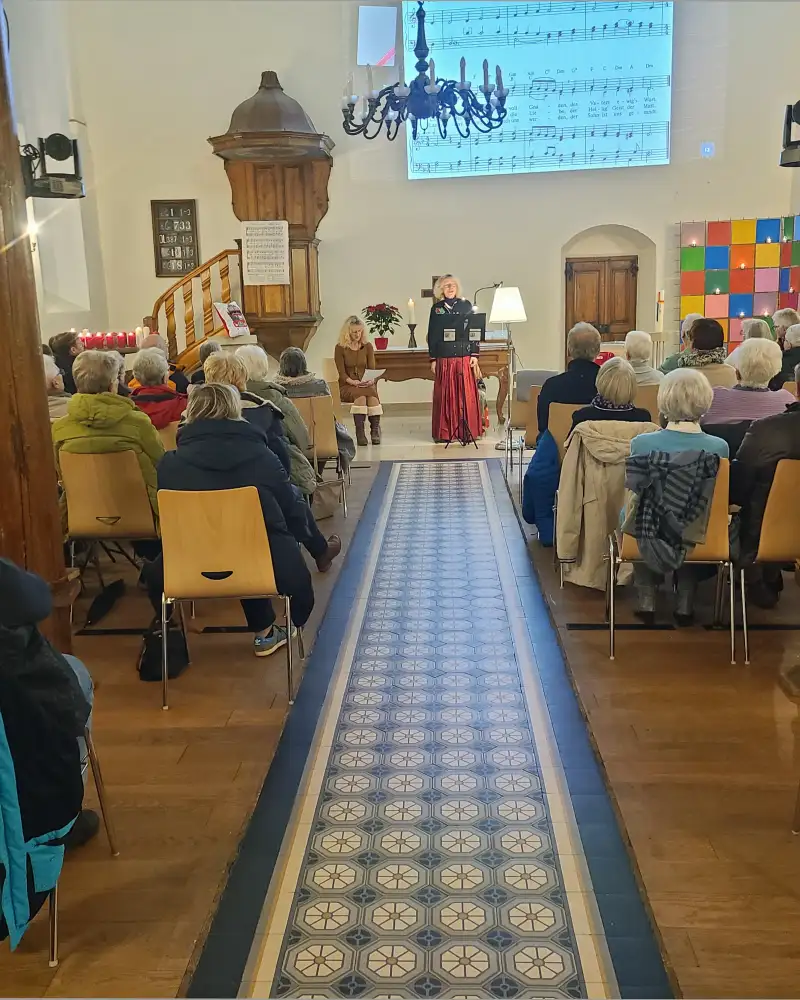 Blick vom Eingang her in einen gut besuchten Kirchenraum. Vorne steht eine Frau in einem langen Rock, vor der ein Pult aufgebaut ist. Neben ihr sitzt eine andere Frau.