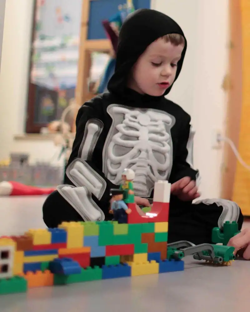 Ein kleiner Junge in einem Kostüm mit aufgedruckten Skelett baut ein Gebäude aus Legosteinen.