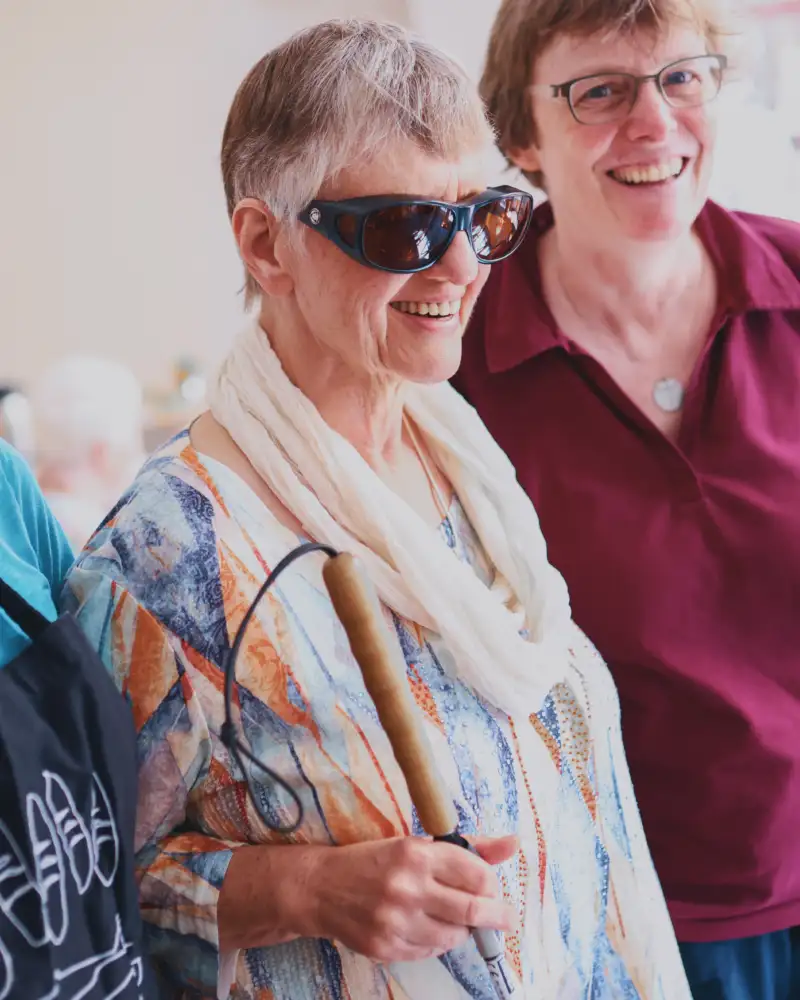 Drei Damen stehen lachend nebeneineiner, eine mit Sonnenbrille und Führstock