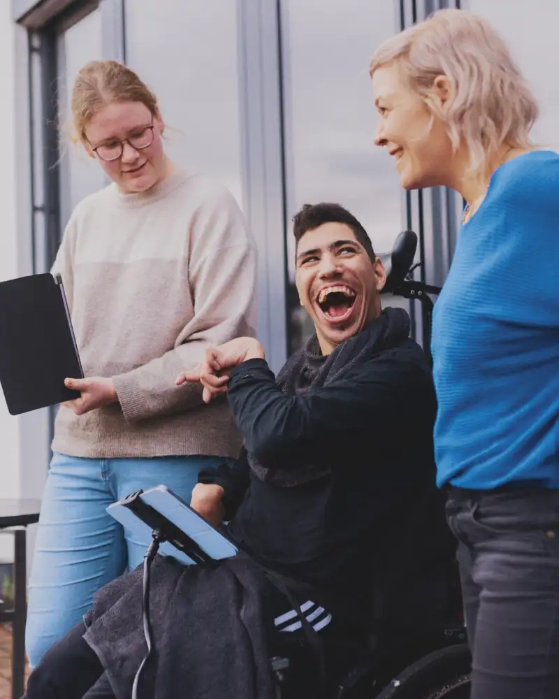 Ein junger Mann im Rollstuhl hat ein Tablet in einer Halterung vor sich und lacht in die Kamera. Rechts und links von ihm stehen zwei Frauen, von denen eine ein Tablet in der Hand hält.