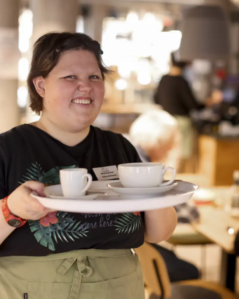 Eine Frau mit Schürze trägt ein Tablett mit Tassen in einem Café.