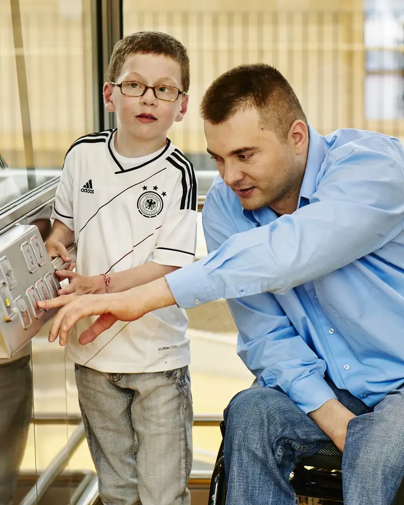 Ein Mann im Rollstuhl, neben ihm ein kleiner Junge. Der Mann bedient einen Fahrstuhl.