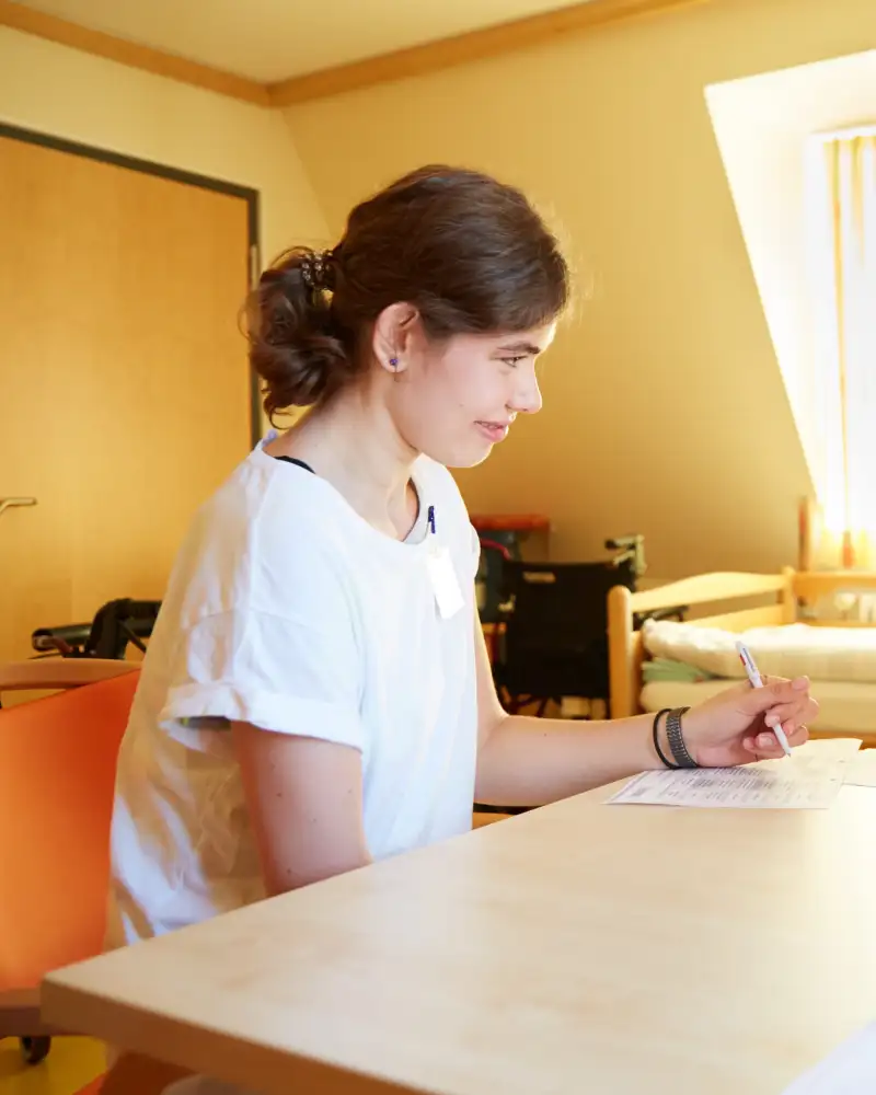 Eine Frau sitzt mit einer weißhaarigen Dame an einem Tisch und schreibt etwas auf ein Formular. Im Hintergrund steht ein Bett.