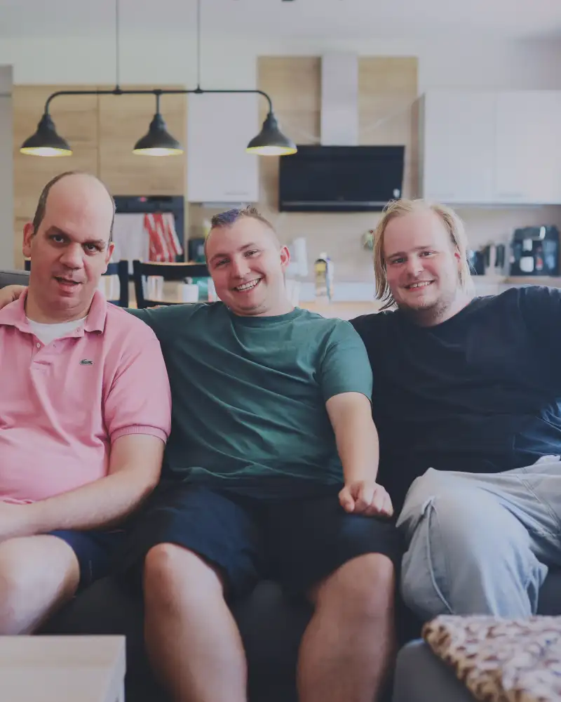 Drei junge Männer sitzen in einem großen Wohnraum nebeneinander auf einem Sofa.