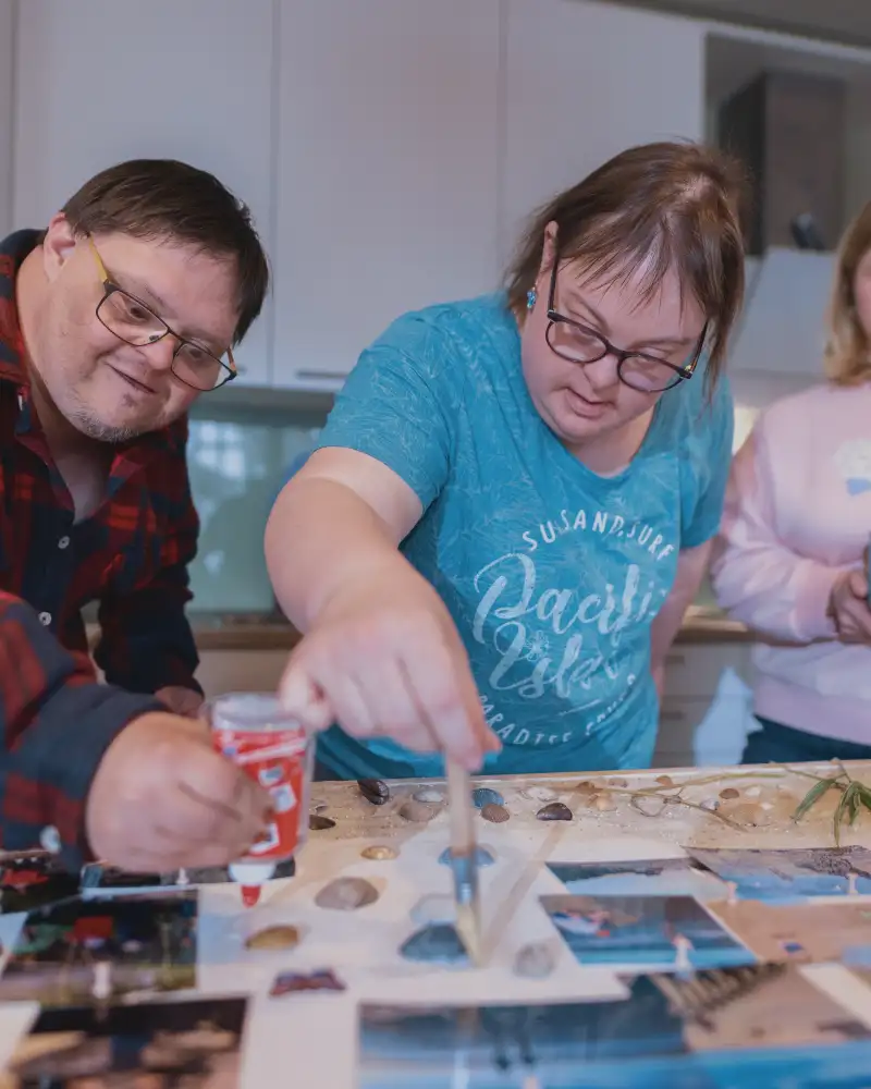 Drei Personen mit Downsyndrom sitzen an einem Tisch, auf dem viele Urlaubsfotos liegen.