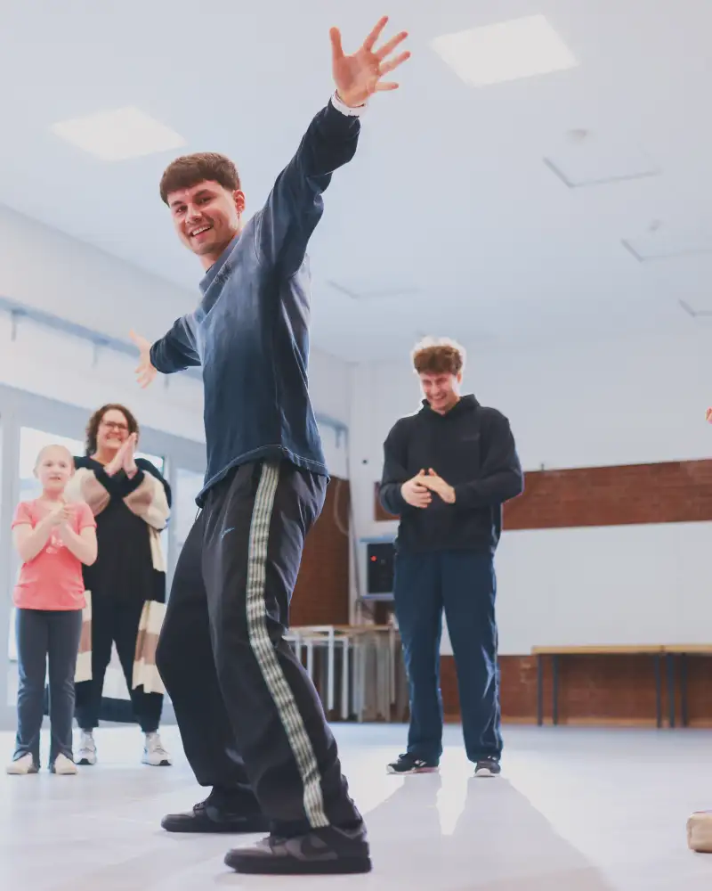 Ein Gruppe unterschiedlichen Alters in einem Übungssaal. Vorne steht ein Jugendlicher mit leicht gebeugten Knien und ausgebreiteten Armen. Die anderen schauen zu.
