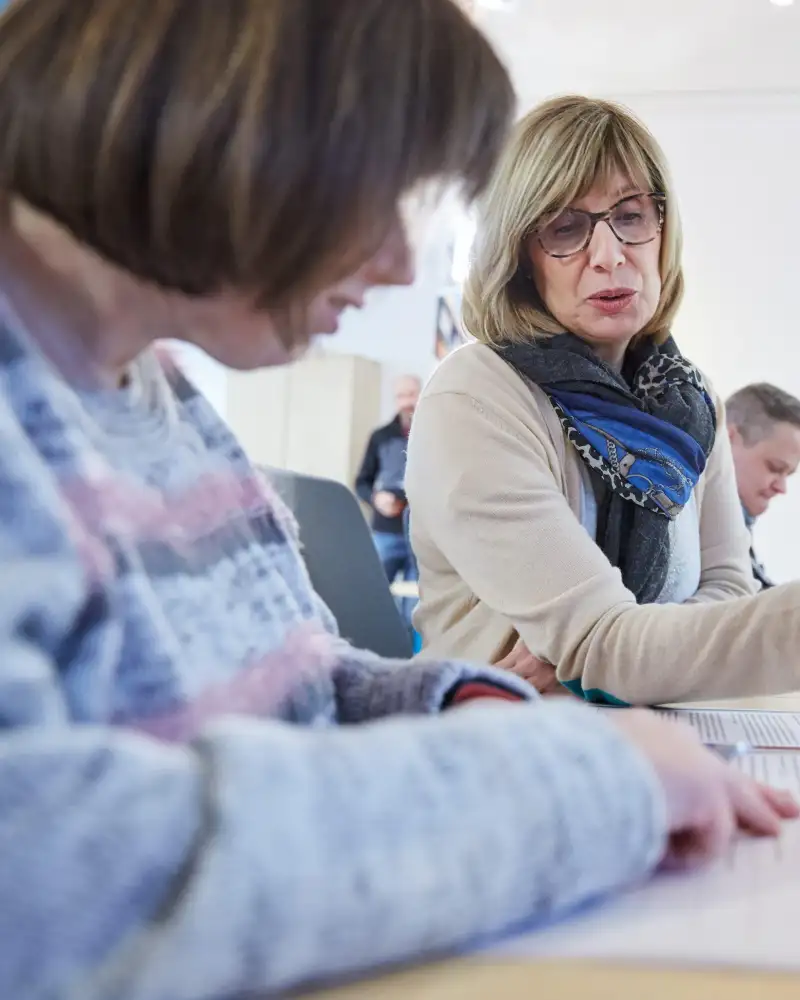 Eine Frau liest in Formularen, die vor ihr liegen. Neben ihr sitzt eine ander Frau, die zu ihr spricht. Im Hintergrund sitzen weitere Personen.
