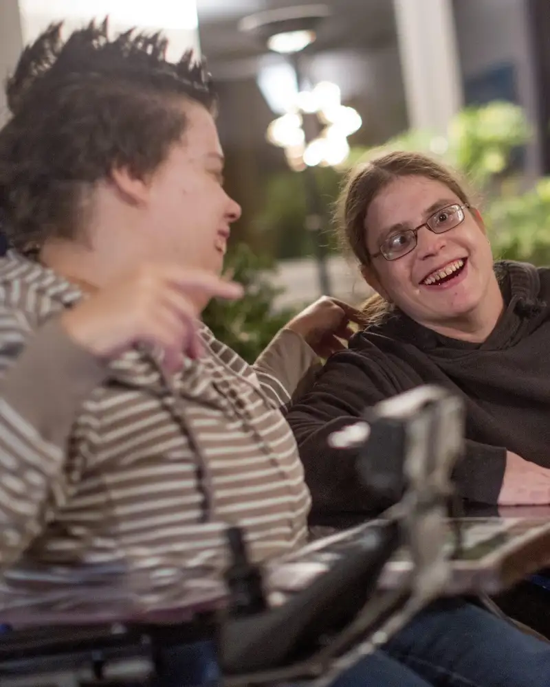 Zwei Frauen sitzen nebeneinander an einem Tisch und lachen zusammen. Eine von ihnen sitzt im Rollstuhl.