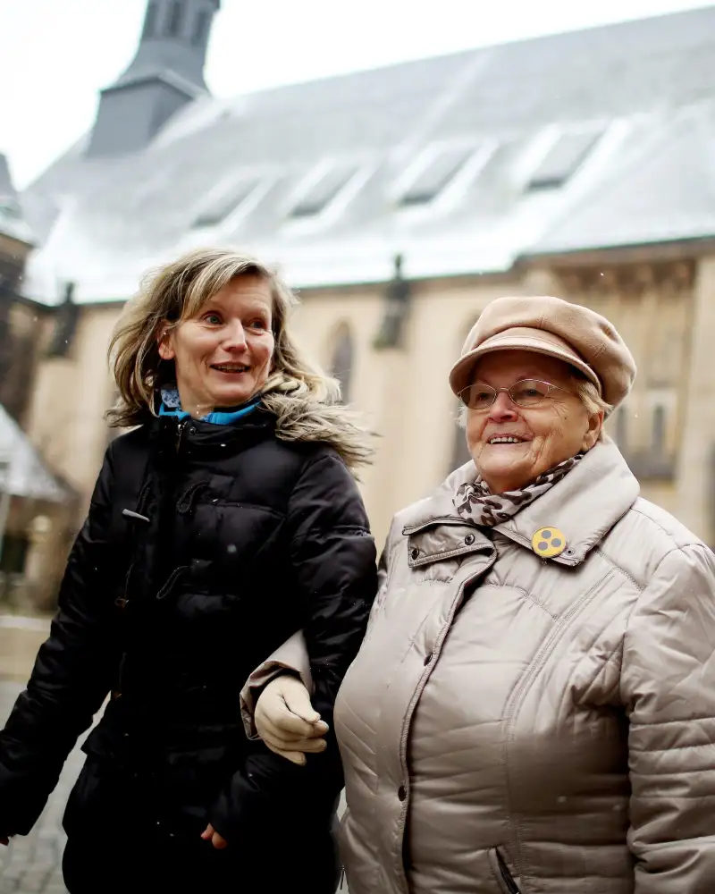 Eine ältere blinde Dame geht an der Hand einer jüngeren Dame durch die Stadt. Im Hingergrund ist eine Kirche zu sehen.