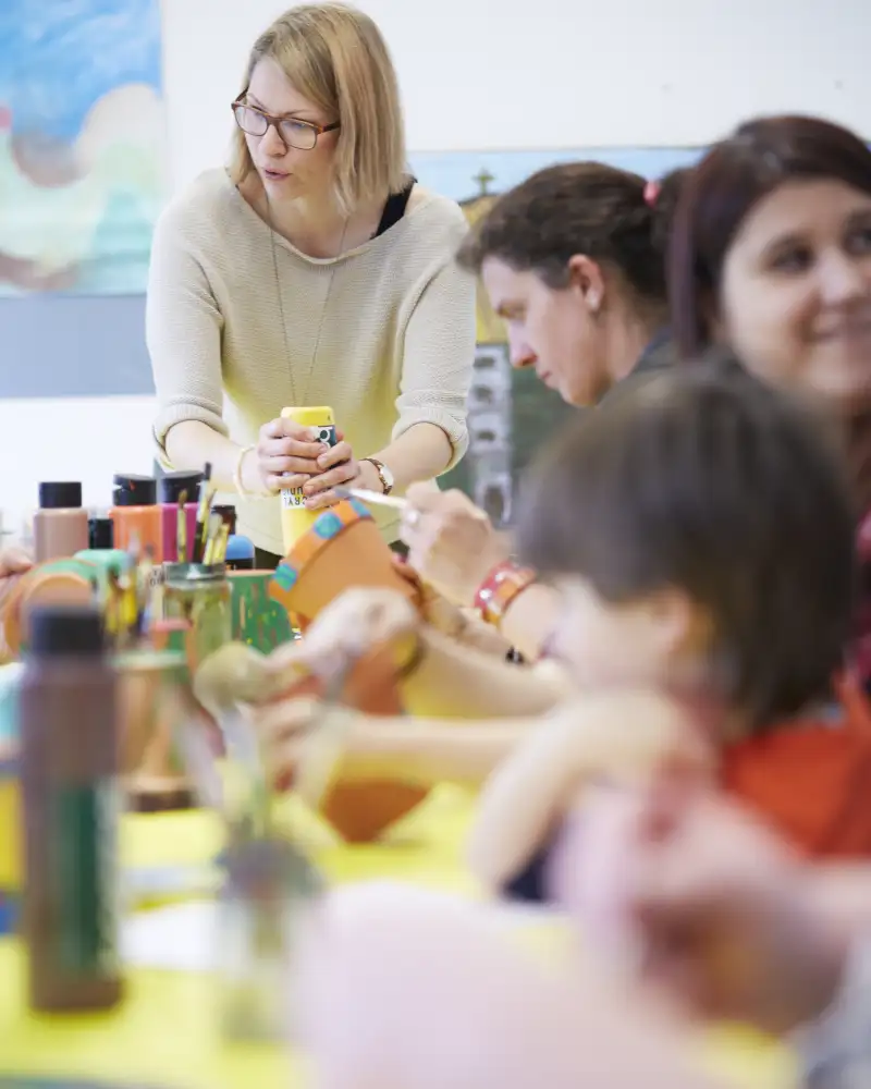 Drei Frauen und ein Kind sitzen mit anderen Personen an einem Tisch. Darauf stehen verschiedene Farbflaschen zum Basteln.