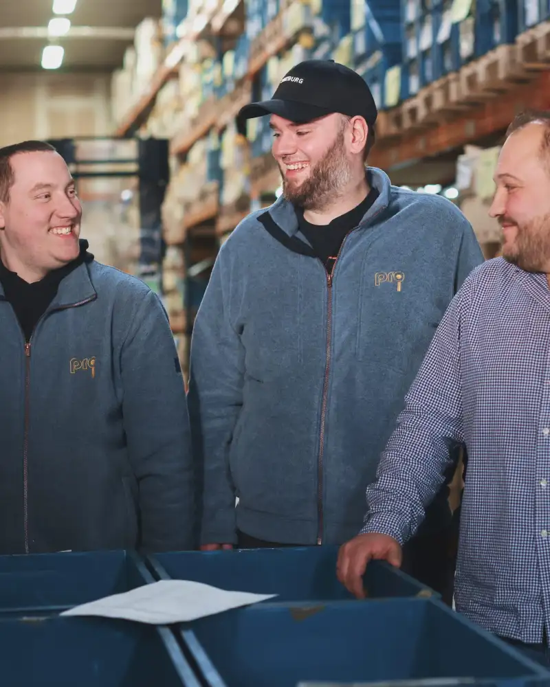 Drei Männer in blauer Kleidung stehen in einem Lagerraum.