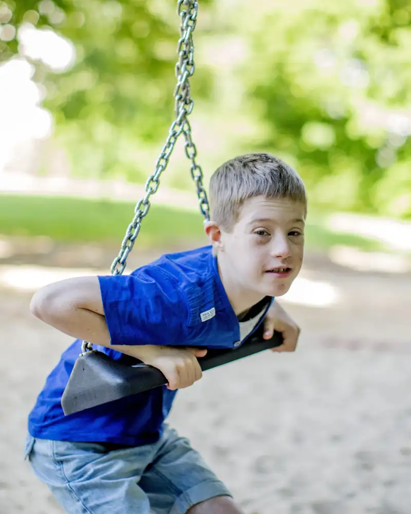 Ein Junge mit geistiger Behinderung auf einem Spielplatz, im Hintergrund Laubbäume. Er hat sich mit dem Oberkörper auf eine Schaukel gelegt.
