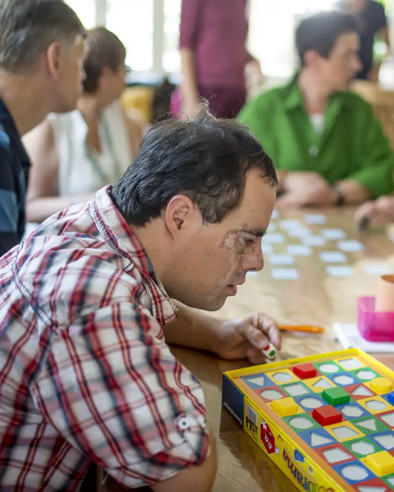 Ein Mann beugt sich über ein Spiel mit kleinen geometrischen Figuren. Am Tisch sitzen weitere Personen und spielen Memory.