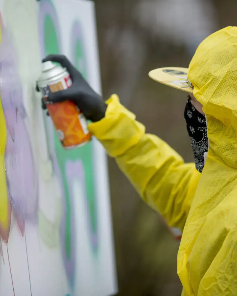 Eine Person im gelben Kapuzenoverall mit einem Tuch vor Mund und Nase besprayt eine weiße Fläche mit bunten Graffiti.