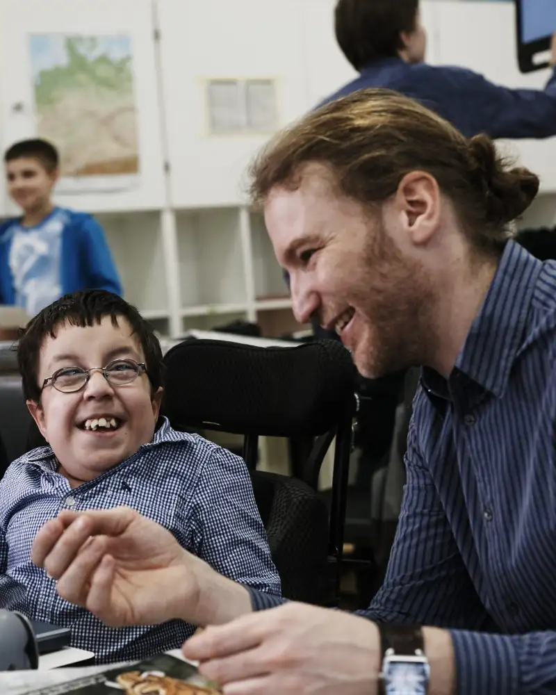 Im Klassenzimmer: ein kleinwüchsiger Schüler spricht mit einem jungen Mann, der neben ihm sitzt.
