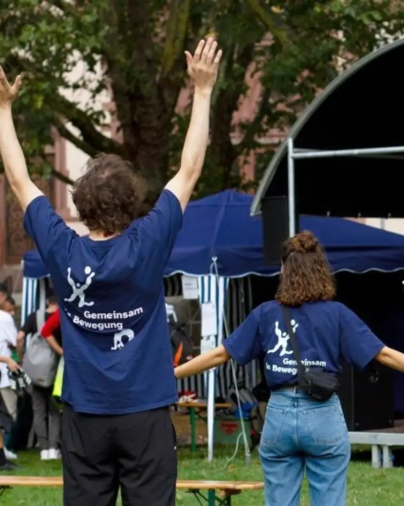 Zwei junge Frauen auf einem Sportplatz. Eine hält beide Arme hoch, die andere breitet ihre Arme aus. Sie tragen blaue T-Shirts mit dem Aufdruck "Gemeinsam in Bewegung".