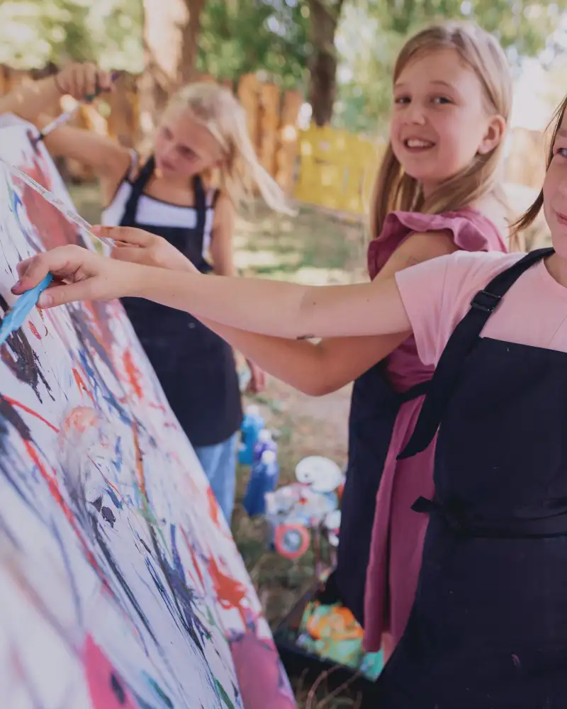 Ein kleines Mädchen malt auf einer Leinwand, neben ihr stehen zwei andere kleine Mädchen.