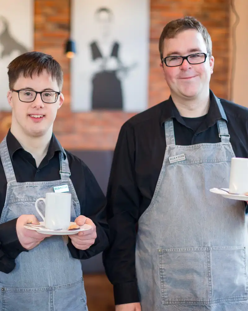 Zwei Männer in einem Restaurant. Sie tragen Schürzen und halten Tassen mit Untertassen in den Händen. Einer von ihnen hat das Downsyndrom.