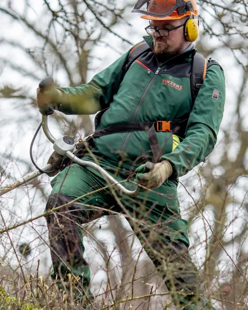 Ein Mann mit Helm und grüner Arbeitskleidung steht mit einem Gartengerät mitten im Gestrüpp.