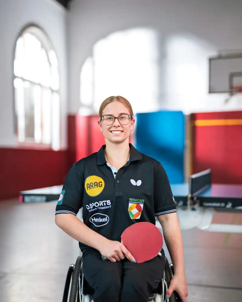 Eine Frau im Rollstuhl mit einem Tischtennisschläger in einer Turnhalle