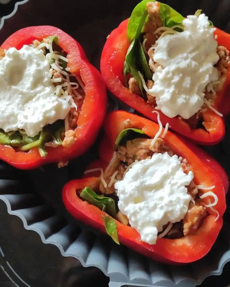 Ein Teller mit drei halbierten roten Paprika. Sie sind jeweils gefüllt mit Salatblatt, Tomatenreis und darauf jeweils ein Klecks Hüttenkäse.