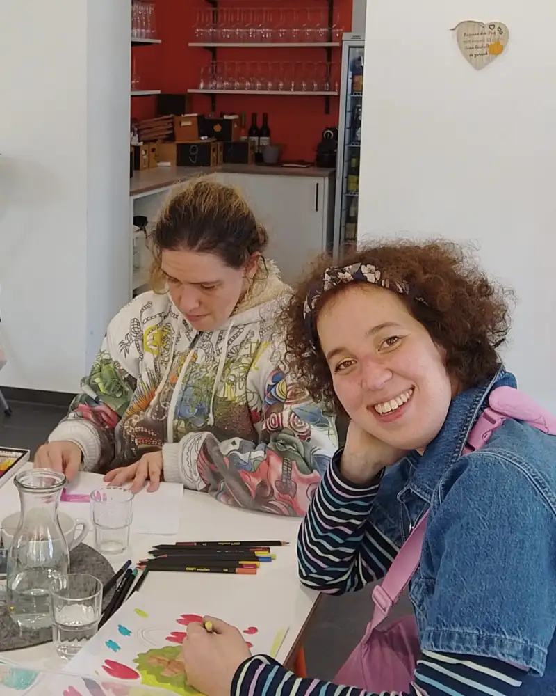 Zwei Frauen sitzen an einem Tisch mit Malutensilien. Eine malt, die andere lächelt in die Kamera.