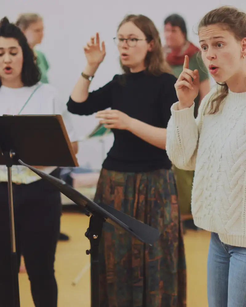 Drei junge Frauen stehen vor Notenpult und singen.